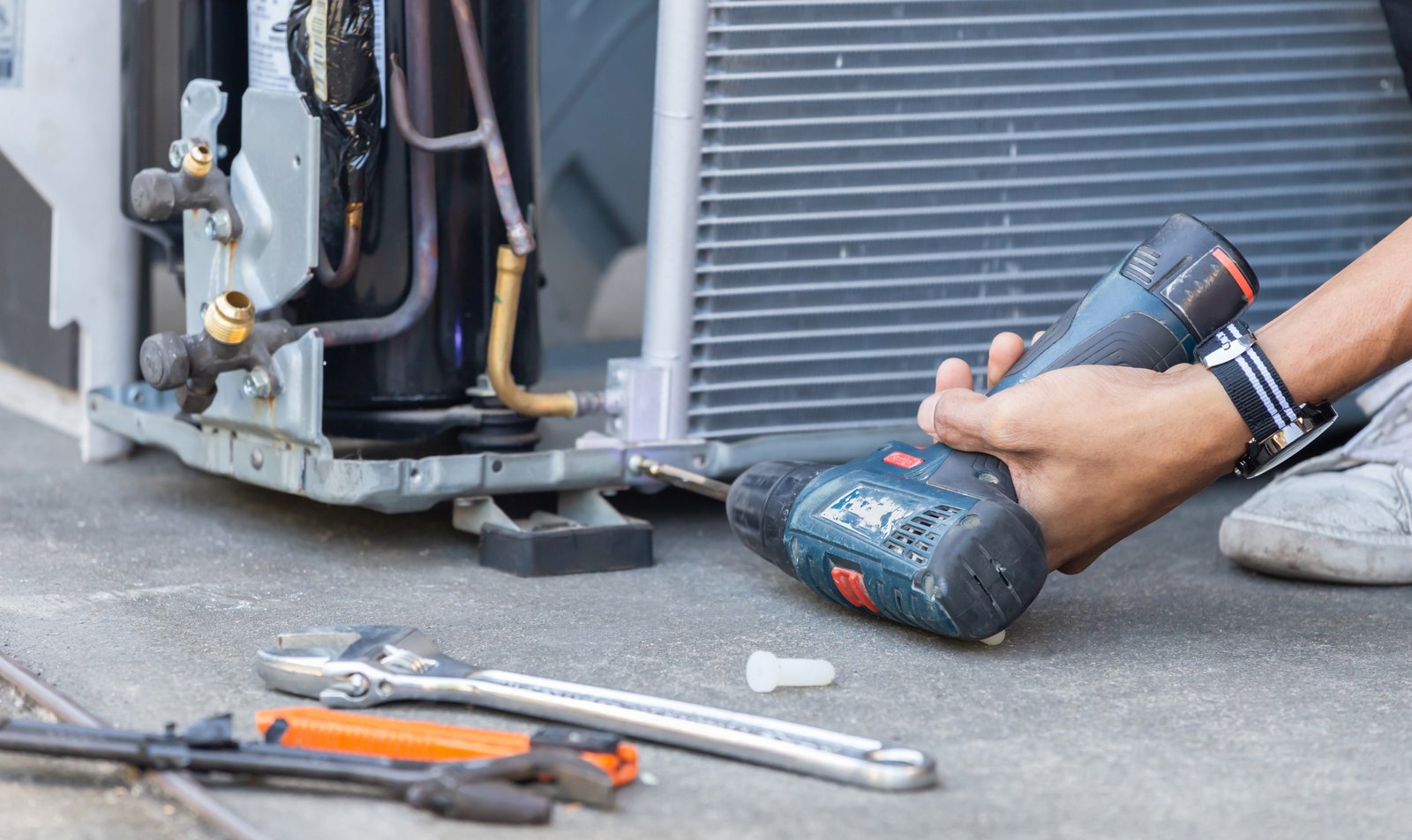 HVAC repair technician using a power drill to fix an air conditioning compressor unit, with tools scattered nearby for repair work.